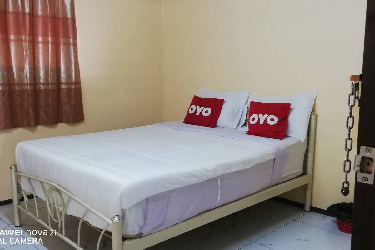 A single-bed room with a white bedspread, two red pillows with 'OYO' branding, a metal bed frame, a red curtain, and a white wall.