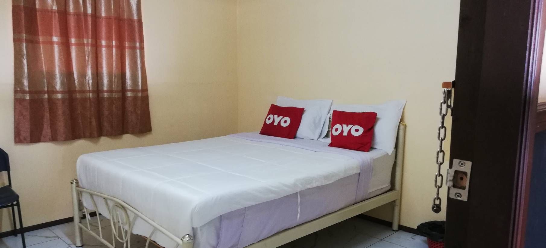 A single-bed room with a white bedspread, two red pillows with 'OYO' branding, a metal bed frame, a red curtain, and a white wall.