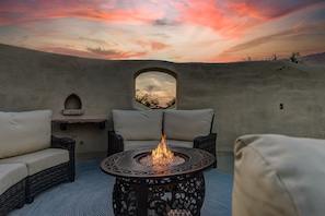 A cozy outdoor seating area with a fire pit, wicker furniture, and a view of the sunset through a window.