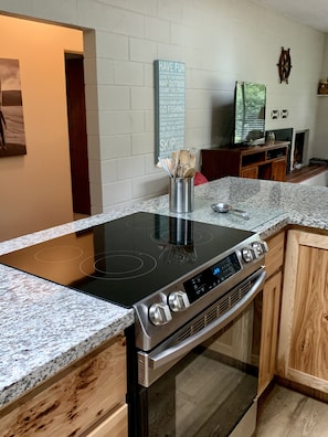 A modern kitchen with a built-in induction cooktop, stainless steel oven, granite countertop, wooden cabinets, and a wall-mounted sign with outdoor activities. - Cozy House in Beautiful Sunny Hiouchi! (Crescent City)