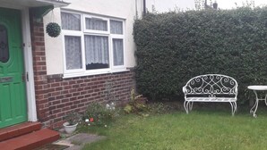 Una casa con una porta verde, facciata in mattoni e una finestra bianca con tende di pizzo. Nel giardino, una panca in metallo bianco e un piccolo tavolo rotondo.