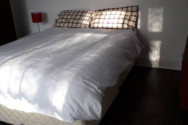 A bed with a white duvet and checkered pillows, a red bedside lamp, and a dark wooden floor.