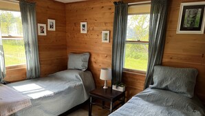 A cozy cabin-style room with two single beds, a small table with a lamp, and framed pictures on the walls. - Cozy cottage, steps from the beach. (Lubec)