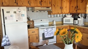 A kitchen with wooden cabinets, a refrigerator, a stove, and a microwave. There is a dining table with a vase of sunflowers. - Cozy cottage, steps from the beach. (Lubec)
