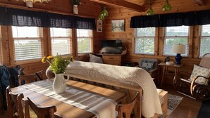 A cozy cabin interior with a wooden dining table, a bench, a television, and a small kitchen area. - Cozy cottage, steps from the beach. (Lubec)
