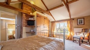 A bedroom with a large bed, wooden beams, a brick fireplace, a television, and a window with curtains. - Church Cottage (CA), Castle Acre, Norfolk (Castle Acre)