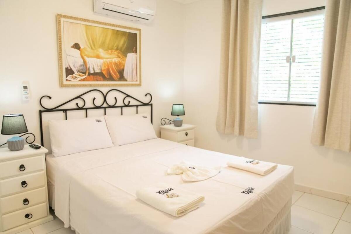 Uma cama bem arrumada com lençóis e toalhas brancas, um abajur, um quadro na parede e uma janela com cortinas.