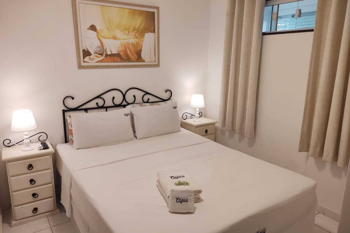 Uma cama bem arrumada com lençóis brancos, duas mesas de cabeceira com abajures, um quadro acima da cama e uma janela com cortinas.