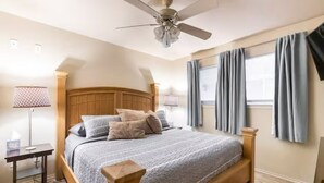 Un dormitorio con una cama de madera, un ventilador de techo, una mesita de noche, una lámpara y una ventana con cortinas.