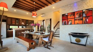 A spacious living area with a wooden dining table, chairs, and a bench. There is a fireplace with a modern design, a kitchen area with a microwave, and a staircase leading to another level. - Alea Villa (Ungasan)