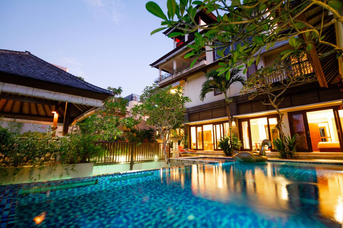 A swimming pool with a tiled edge, surrounded by lush greenery and a building with a balcony.