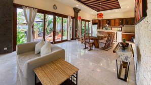 A spacious living area with a sofa, dining table, and chairs, a kitchen with wooden cabinets, and large sliding glass doors leading to a balcony with outdoor furniture. - Alea Villa (Ungasan)