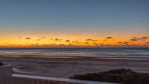 Vista del atardecer desde un balcón con vista a una playa, cielo despejado y luces de la ciudad a lo lejos.
