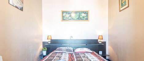 Hotellihuone, jossa on sänky, yöpöytävalaisimet, seinälle ripustettu taideteos ja pieni pöytä, jonka päällä on kasvi.