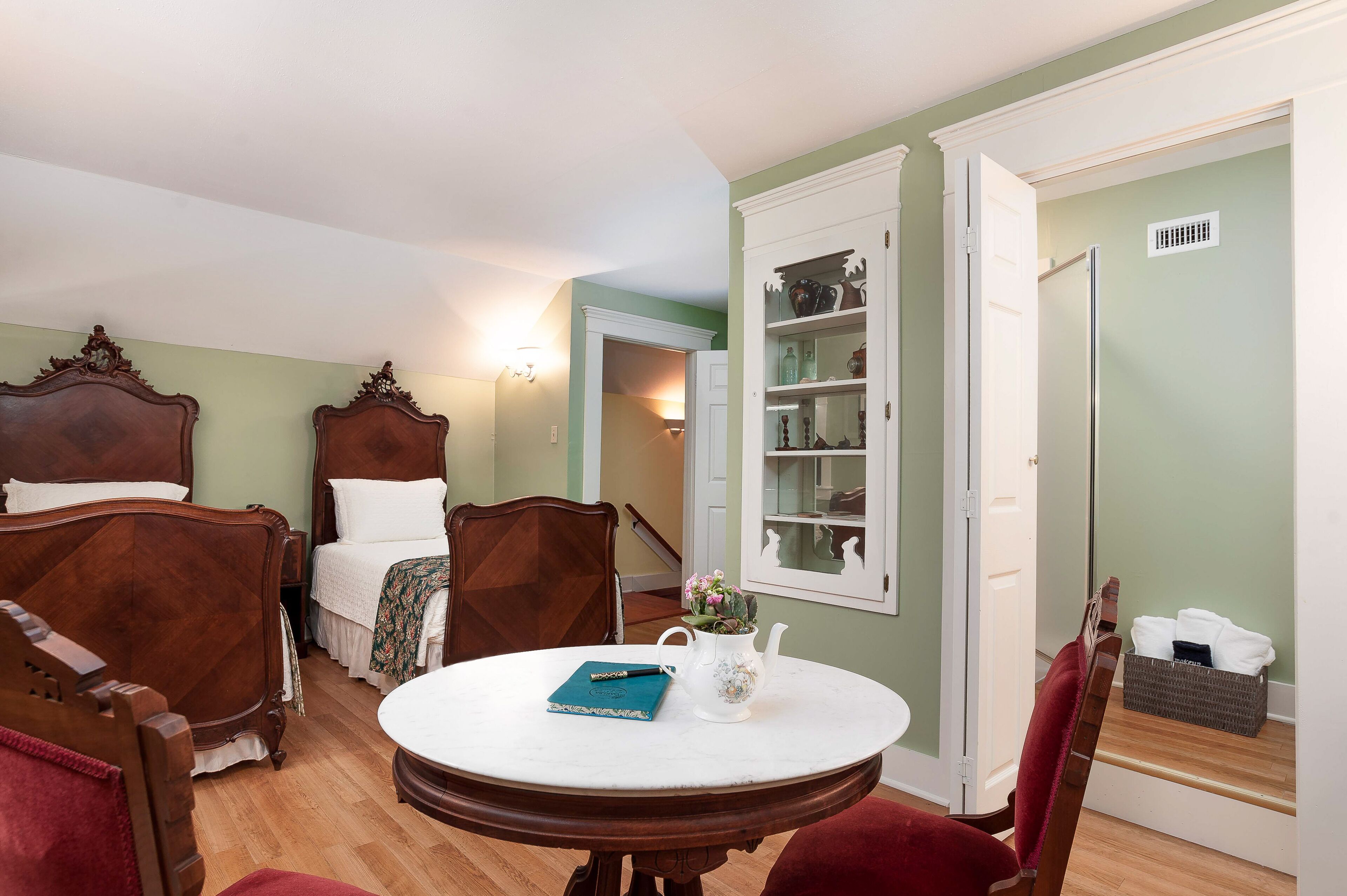 A bedroom with a bed, two chairs, a round table with a book and a teapot, and a wardrobe with shelves.