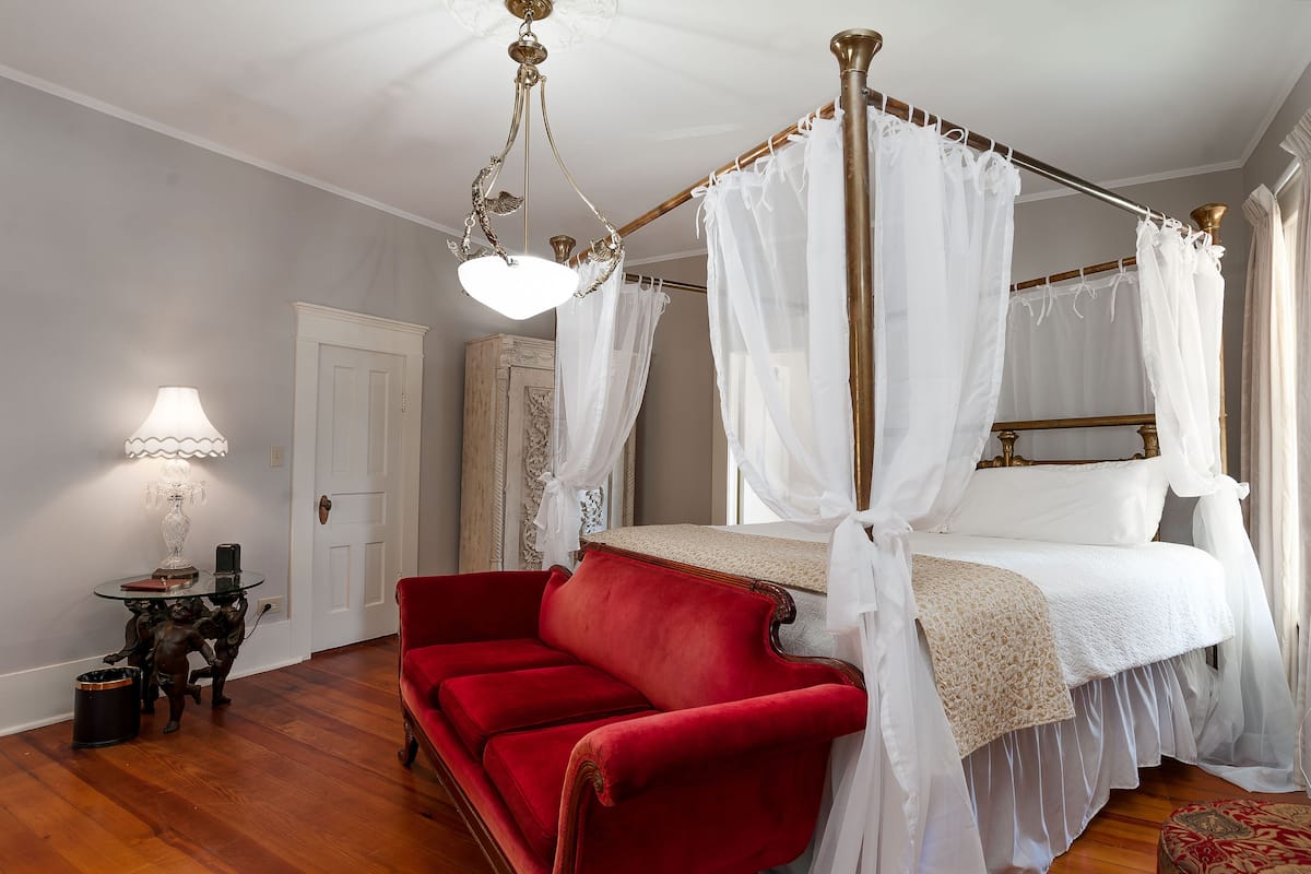 Un lit à baldaquin à quatre poteaux avec une literie blanche, un fauteuil en velours rouge, une petite table avec une lampe et un plancher en bois.