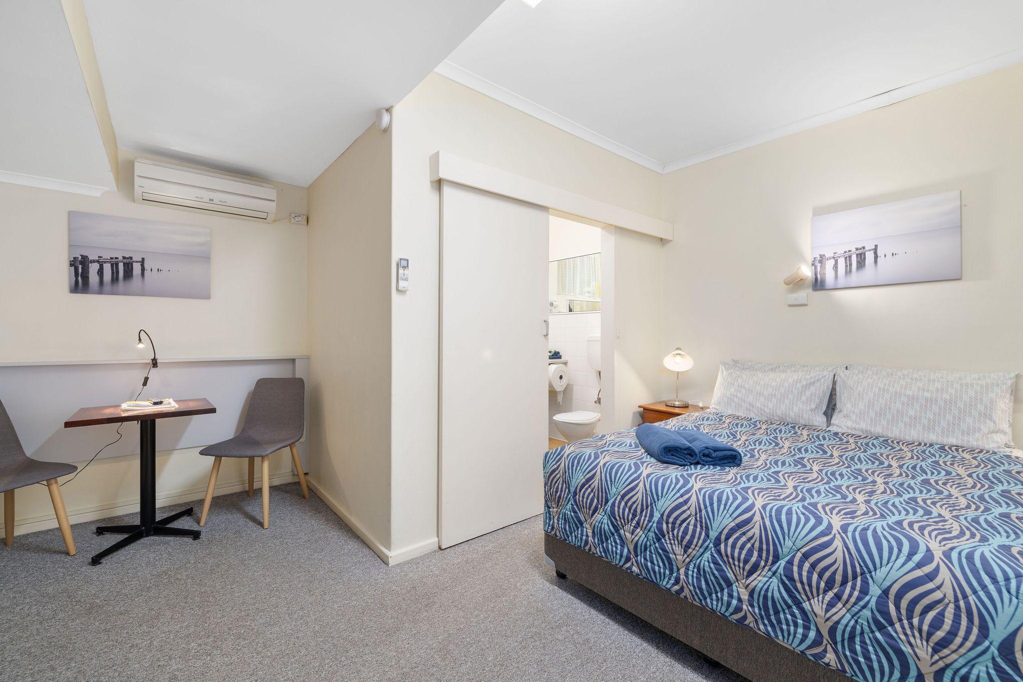 A hotel room with a bed, a desk with two chairs, a door, and a wall-mounted air conditioner.