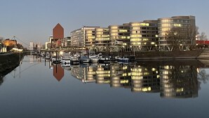 A waterfront cityscape with modern buildings, a calm river, and boats docked along the shore. - Hausboot Lore (Duisburg)