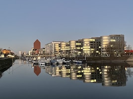 Un paisaje urbano frente al agua con edificios modernos, un río tranquilo y barcos amarrados a la orilla.