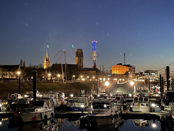 Una marina al anochecer con barcos amarrados, una torre destacada con luces azules y un edificio con una torre de reloj al fondo.