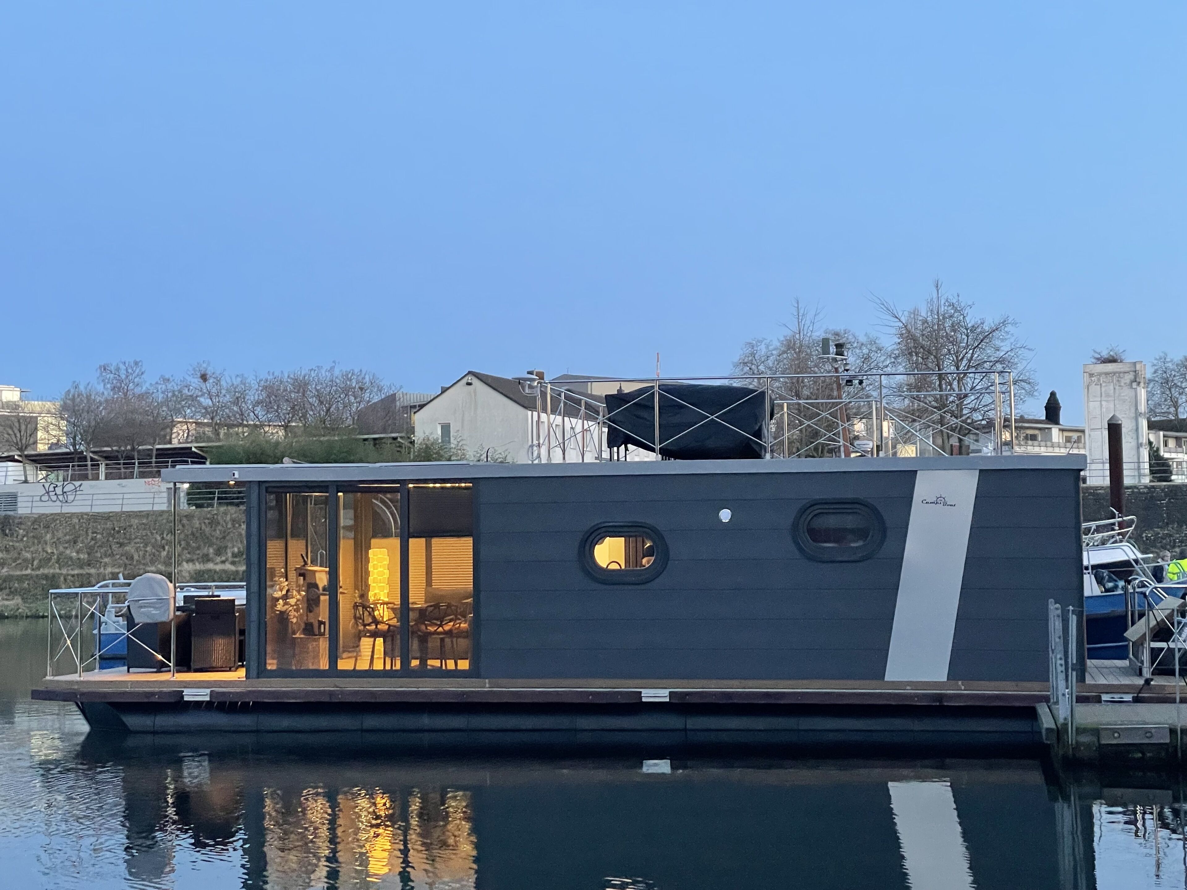 Une maison flottante moderne avec une entrée dotée d’une porte en verre, une petite terrasse et une vue sur l’eau.