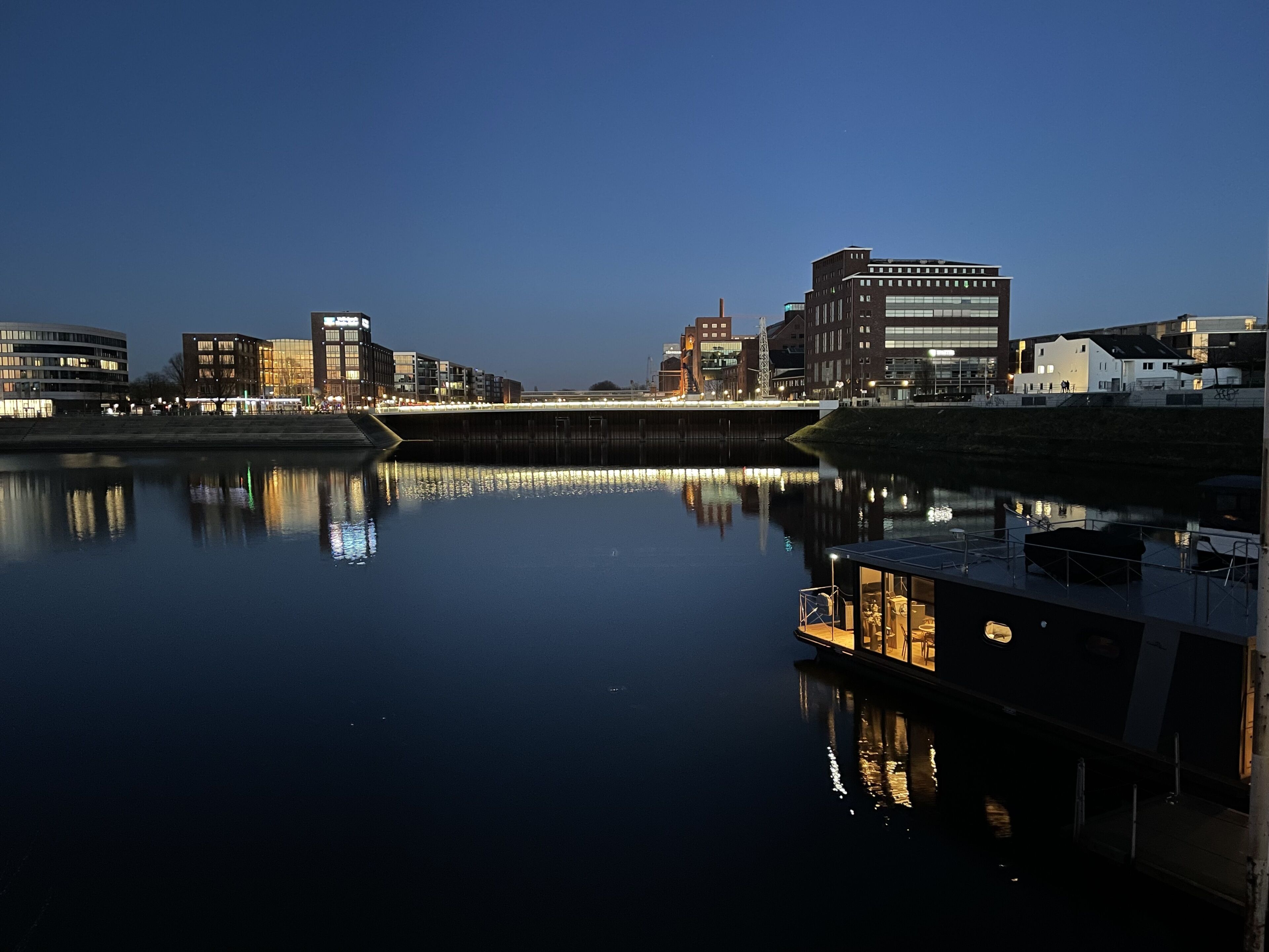 A waterfront view with buildings reflecting on calm water, a dock with a boat, and a bridge in the background.