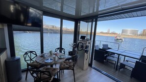 A modern dining area with a view of a river, a boat in the distance, and a glass-enclosed space. - Hausboot Lore (Duisburg)