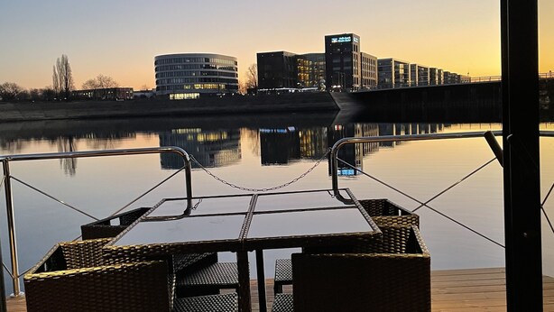 An outdoor deck with a table and chairs, overlooking a calm body of water and buildings in the distance. - Hausboot Lore (Duisburg)