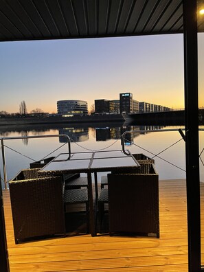 Una terraza al aire libre con una mesa y sillas, con vistas a un cuerpo de agua tranquilo y edificios a lo lejos.