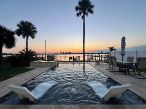 A swimming pool with a view of the city skyline at sunset, surrounded by palm trees and outdoor furniture. - Riverfront Retreat - Pool & Hot Tub - Mins from Beach, Speedway, & Ocean Center! (Daytona Beach)