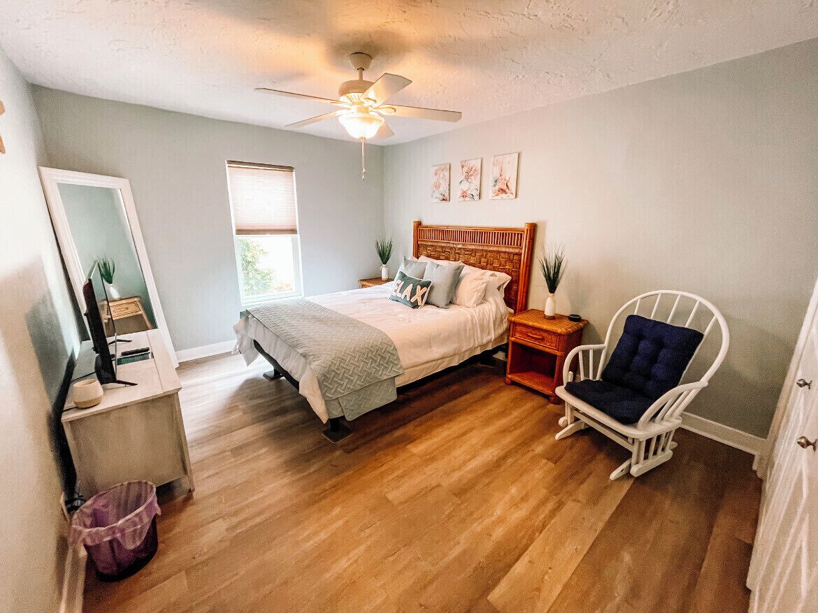 Une chambre à coucher comprenant un lit, une chaise, une table de chevet, un miroir et un ventilateur de plafond.