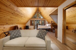 Ein gemütliches Schlafzimmer unter dem Dach mit Holzbalken, einer Couch mit gemusterten Kissen und einem Bett mit Blick ins Freie.