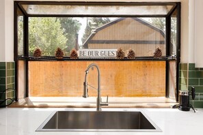 A kitchen with a stainless steel sink, a window with a view of a wooden building, and pine cones on the window sill. - 🐻FamilyCabin🍿TheaterRoom🎱GameRoom🔋EVCharger 🐶PetFriendly (Big Bear)