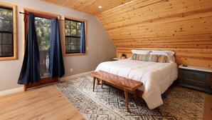 Un dormitorio con techo inclinado, una cama con ropa de cama blanca, un banco, una mesita de noche y una ventana con cortinas.
