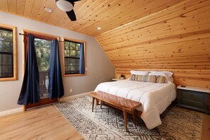 Un dormitorio con techo inclinado, una cama con ropa de cama blanca, un banco, una mesita de noche y una ventana con cortinas.