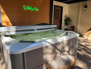 Un bain à remous de spa doté d’un panneau de commande, installé sur une terrasse avec un mur en bois et une enseigne au néon en arrière-plan.