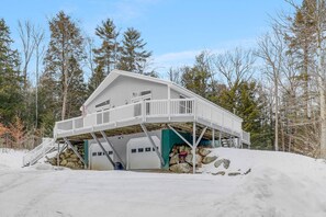 A cabin with a covered deck and two garage doors, surrounded by snow and trees. - Cardinals Perch (Campton)