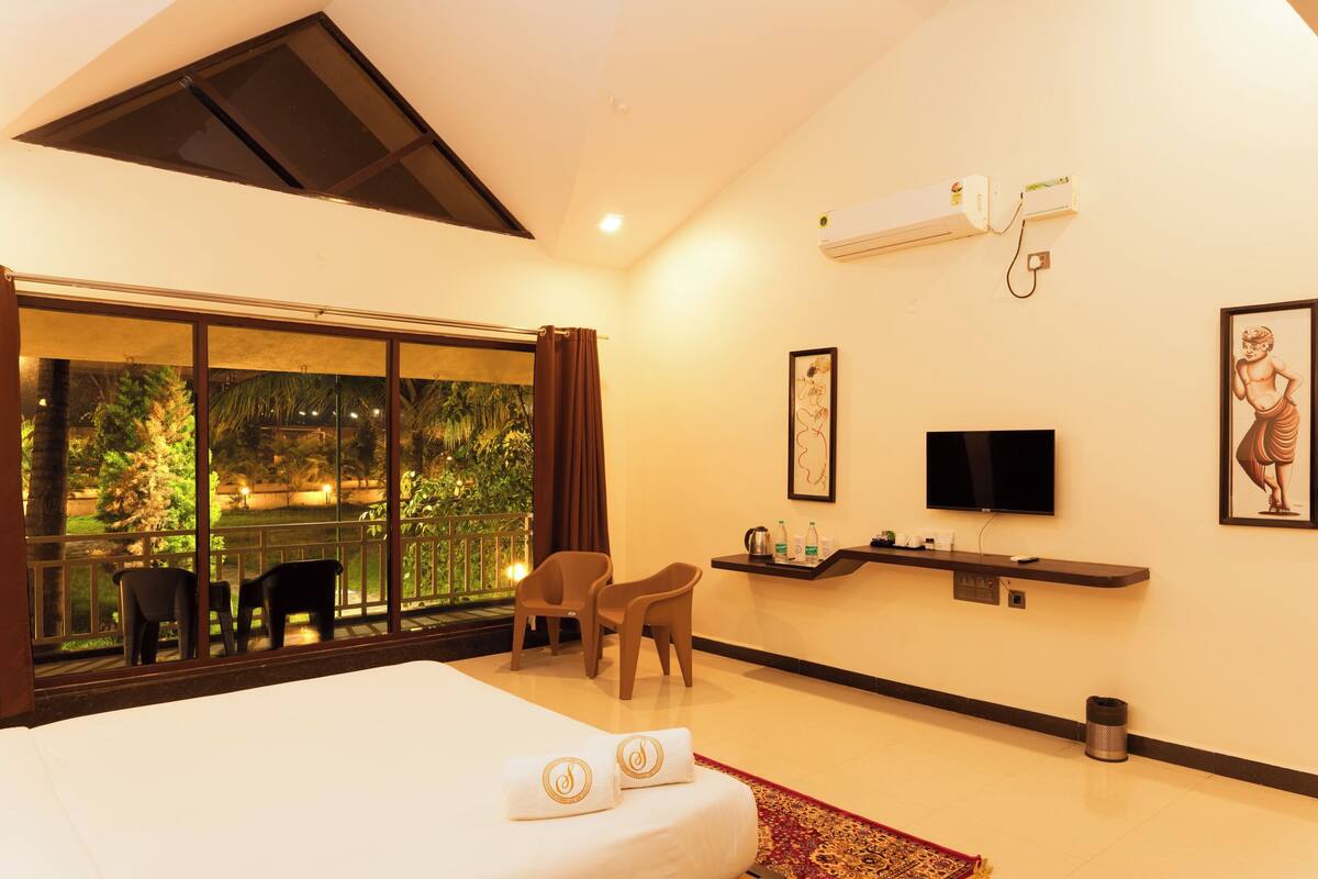 A hotel room with a bed, a TV, a desk with a computer, a window with a view of greenery, and a ceiling fan.