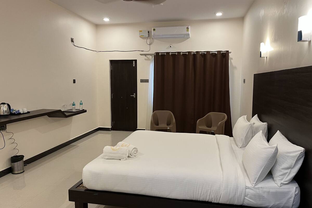A hotel room with a bed, two chairs, a desk with a coffee maker, and an air conditioning unit.