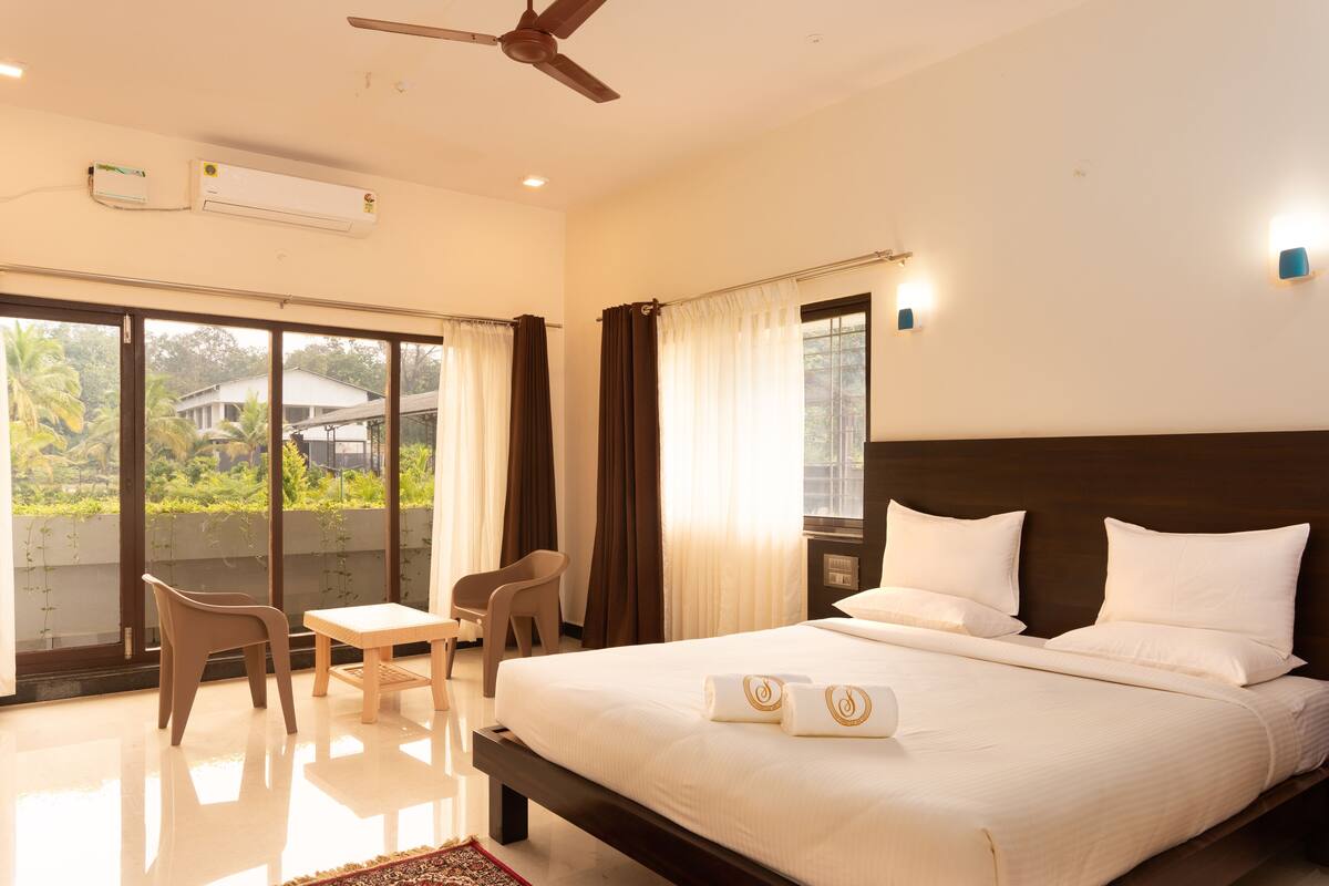 A hotel room with a large bed, a ceiling fan, an air conditioning unit, a small table with a chair, and a view of greenery outside.