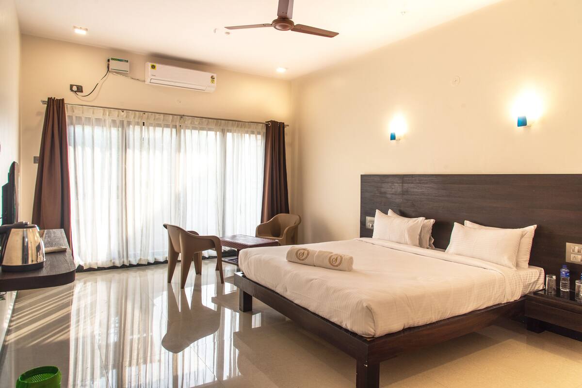A hotel room with a bed, a desk, a chair, a table, a ceiling fan, and an air conditioning unit.