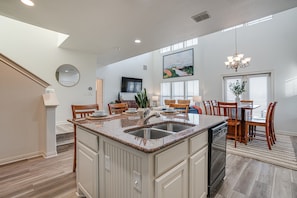 A spacious kitchen with a large island, wooden dining table, and chairs. There is a chandelier, a large painting, and a potted plant.