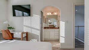 Un dormitorio con cama, escritorio y televisor. Hay un baño con lavamanos y espejo.