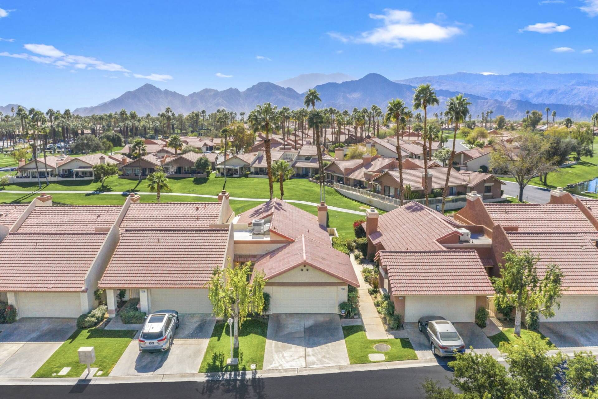 A residential area with houses featuring red-tiled roofs, arranged in rows with green lawns and palm trees.
