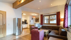 Een moderne woonkamer met een bank, poef en een rode fauteuil. In de achtergrond is een keuken te zien met een koelkast en een magnetron.