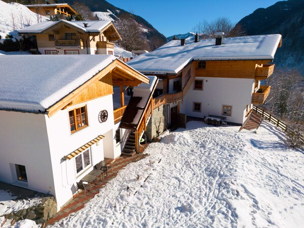 A snow-covered mountain lodge with multiple wooden buildings, a staircase leading to a balcony, and a clear view of the surrounding mountains.