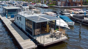 Een drijvend woonschip met dek en overdekt terras, afgemeerd in een jachthaven met andere boten.