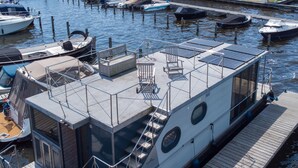 Een drijvend woonschip aan een jachthaven, met een overdekt dek, een klein terras en een trap naar een hoger dek.