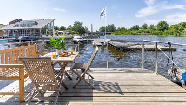 Een houten terras met een ontbijttafel, uitkijkend op een jachthaven met boten en een vlag.