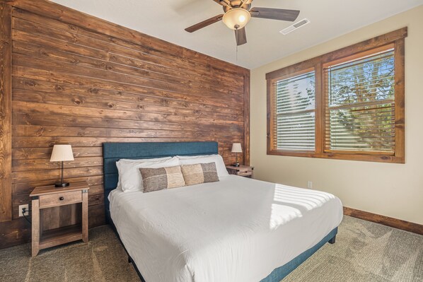 A bedroom with a wooden wall, a bed with white bedding, a ceiling fan, and a window with blinds. - Zion Cabin Retreat - Luxury with Nature Trails in Uncrowded East Zion. 3 kings (Orderville)